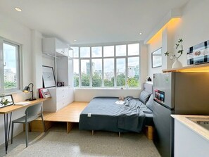 Quarto compacto com cama, escrivaninha e pequeno espaço de cozinha.