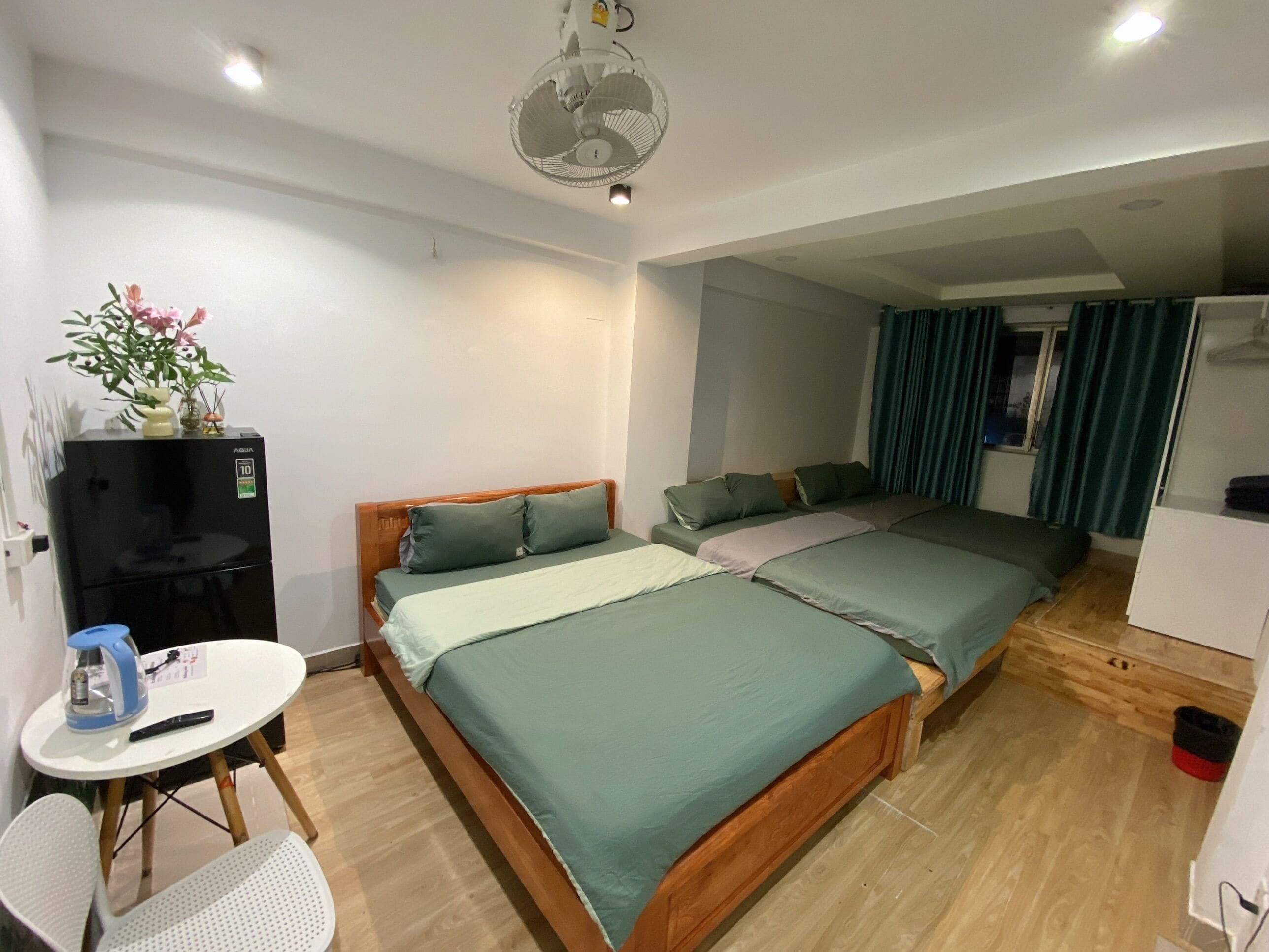 A room with two beds, a small table with a chair, a refrigerator, and a ceiling fan.