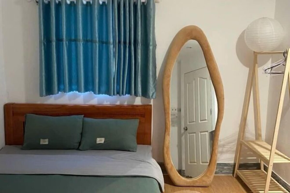 A bed with a wooden headboard, two pillows, a mirror with a wooden frame, a white door, and a wooden shelf with a white spherical object.