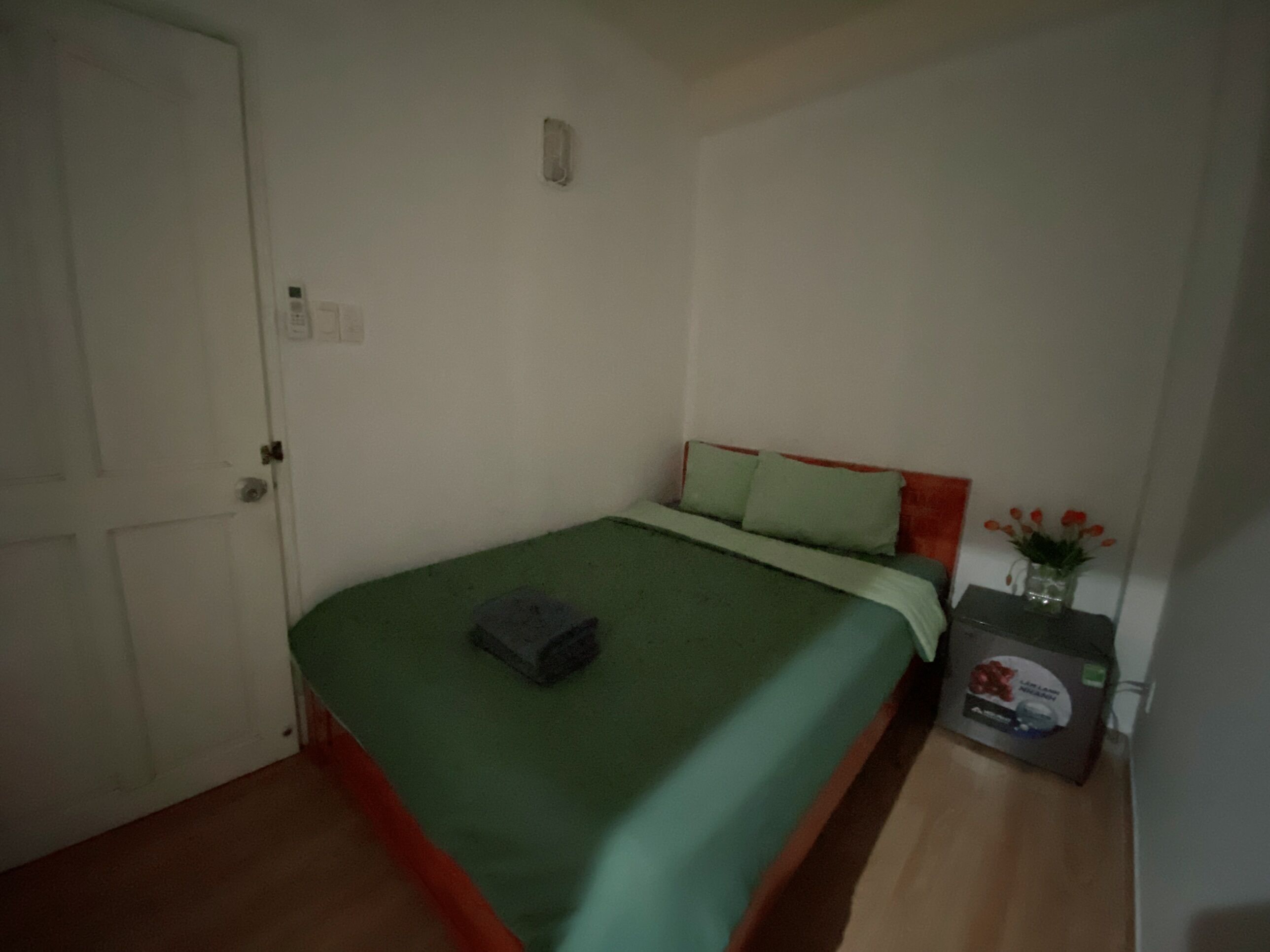 Une petite pièce simple, avec un lit simple, une table de chevet, une porte et une petite table sur laquelle est posé un vase.
