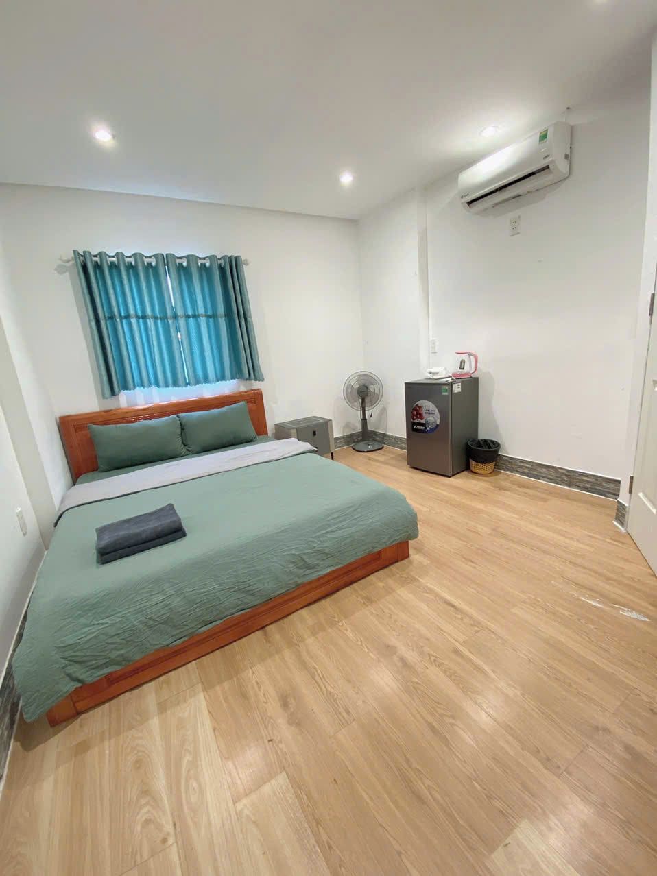 A hotel room with a bed, a fan, an air conditioner, and a small table with a kettle.