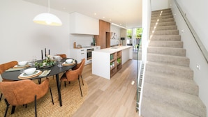 Una cocina moderna con una isla central, un comedor con sillas marrones y una escalera que sube a un piso superior.