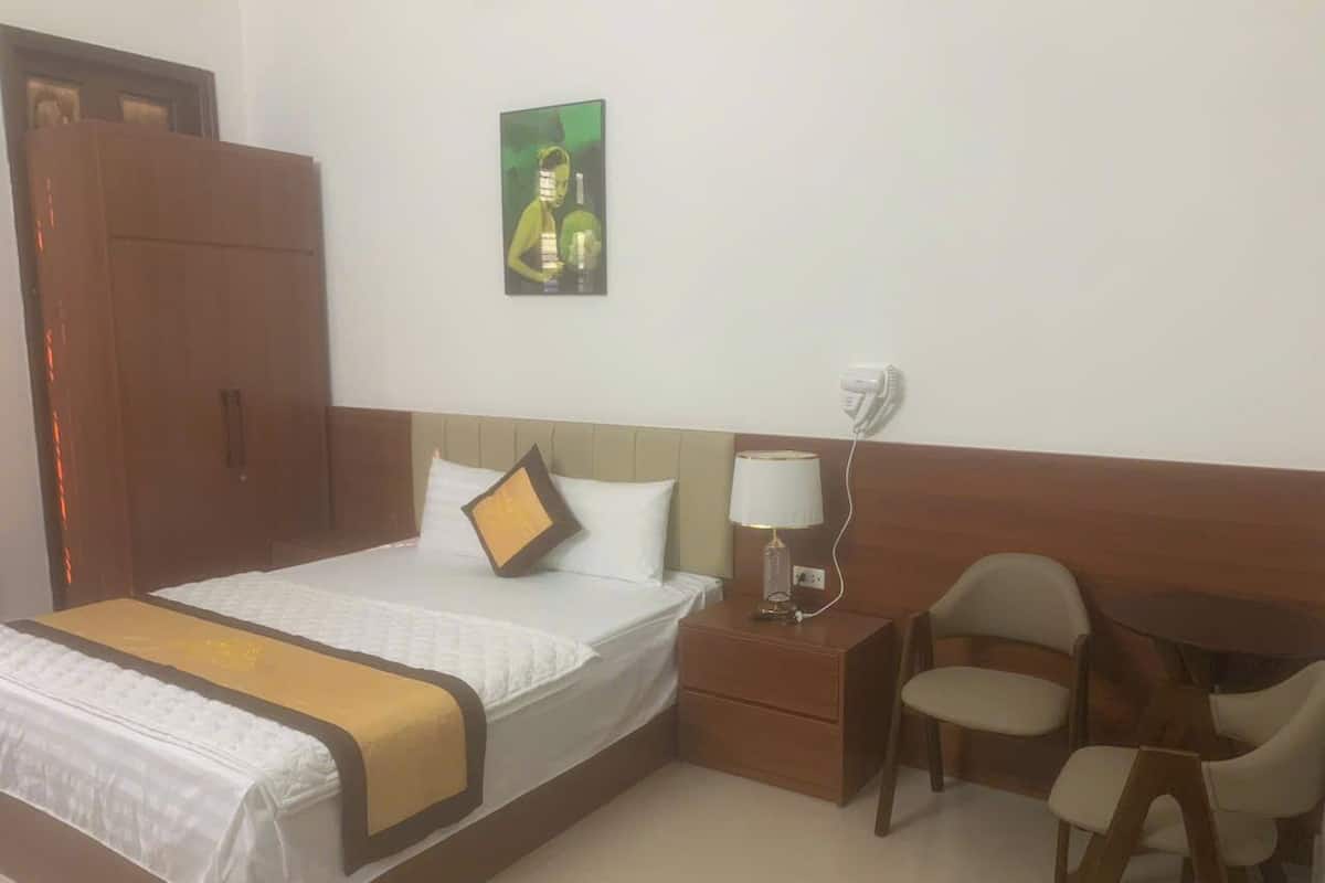 A hotel room with a large bed, a bedside table with a lamp, a chair, and a small table.