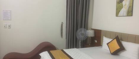 Quarto de hotel com cama, escrivaninha, cadeira, ventoinha e um quadro na parede.