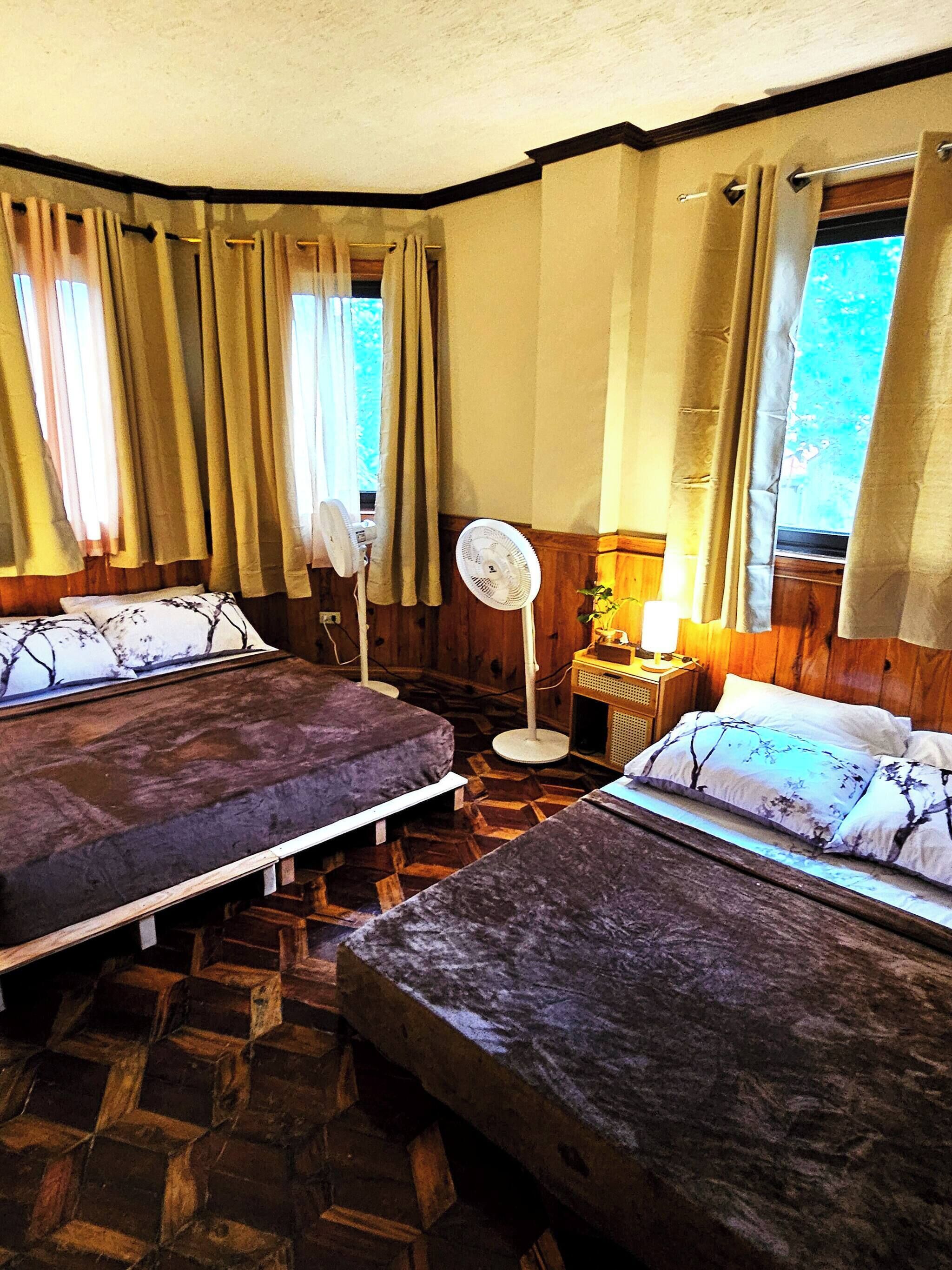 A room with two beds, a standing fan, a nightstand with a lamp, and curtains on the windows.