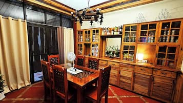 A traditional wooden dining area with a table and chairs, a fan, and a kitchen area with wooden cabinets and shelves.