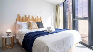 A neatly made bed with white linens, a blue throw blanket, and rolled towels on the side. There is a small bedside table with a lamp and a wooden headboard.
