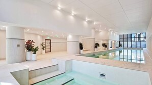 A modern indoor swimming pool area with a glass barrier, seating, and potted plants.