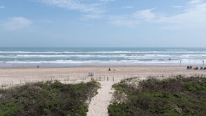 A beach with a sandy path leading to the ocean, a few beachgoers, and beach chairs in the distance.