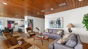 A spacious living room with a wooden ceiling, a gray sofa, wicker chairs, and a dining area with a Texas flag.