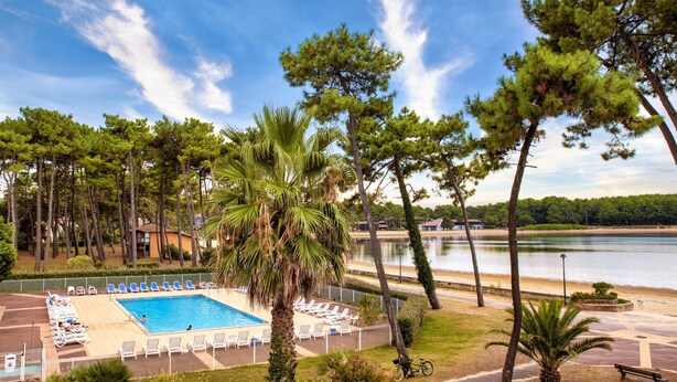 A swimming pool with lounge chairs, surrounded by palm trees and pine trees, with a view of a lake and buildings in the background. - Sowell Family Pignada Plage by Interhome (Soustons)
