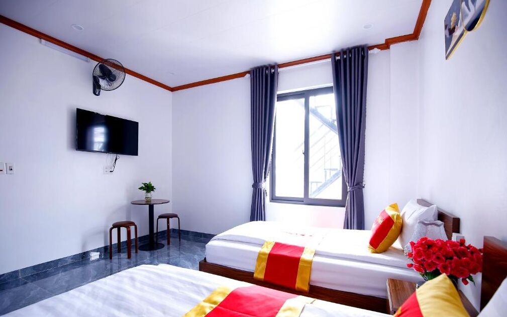 Une chambre d’hôtel avec deux lits, une télévision, une petite table avec une plante et une fenêtre avec des rideaux.