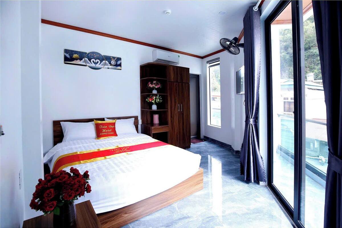 Una habitación de hotel con una cama, un armario de madera con flores y un amplio ventanal con cortinas.