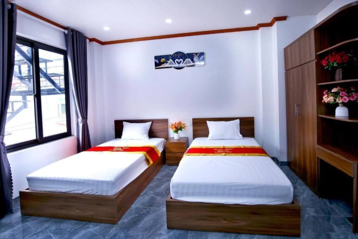 Habitación de hotel con dos camas individuales, un armario de madera, una mesita de noche con un jarrón de flores y una ventana con cortinas.