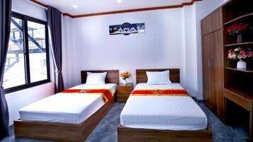 A hotel room with two single beds, a wooden wardrobe, a nightstand with a vase of flowers, and a window with curtains.