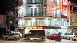 Un immeuble à plusieurs étages, avec une enseigne indiquant « HÔTEL TIEN DAT » au niveau du rez-de-chaussée. Plusieurs véhicules sont stationnés devant, notamment un fourgon et des voitures.
