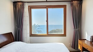 A hotel room with a large bed, a wooden desk, and a view of the sea through a window. - Tteuribada Pension (Seogwipo)