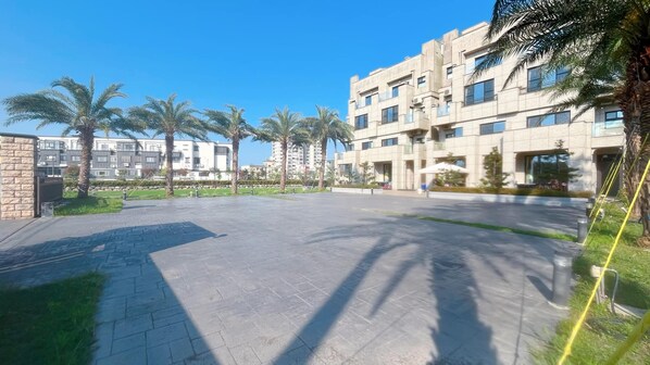 A modern residential building with a paved courtyard, palm trees, and a clear blue sky.