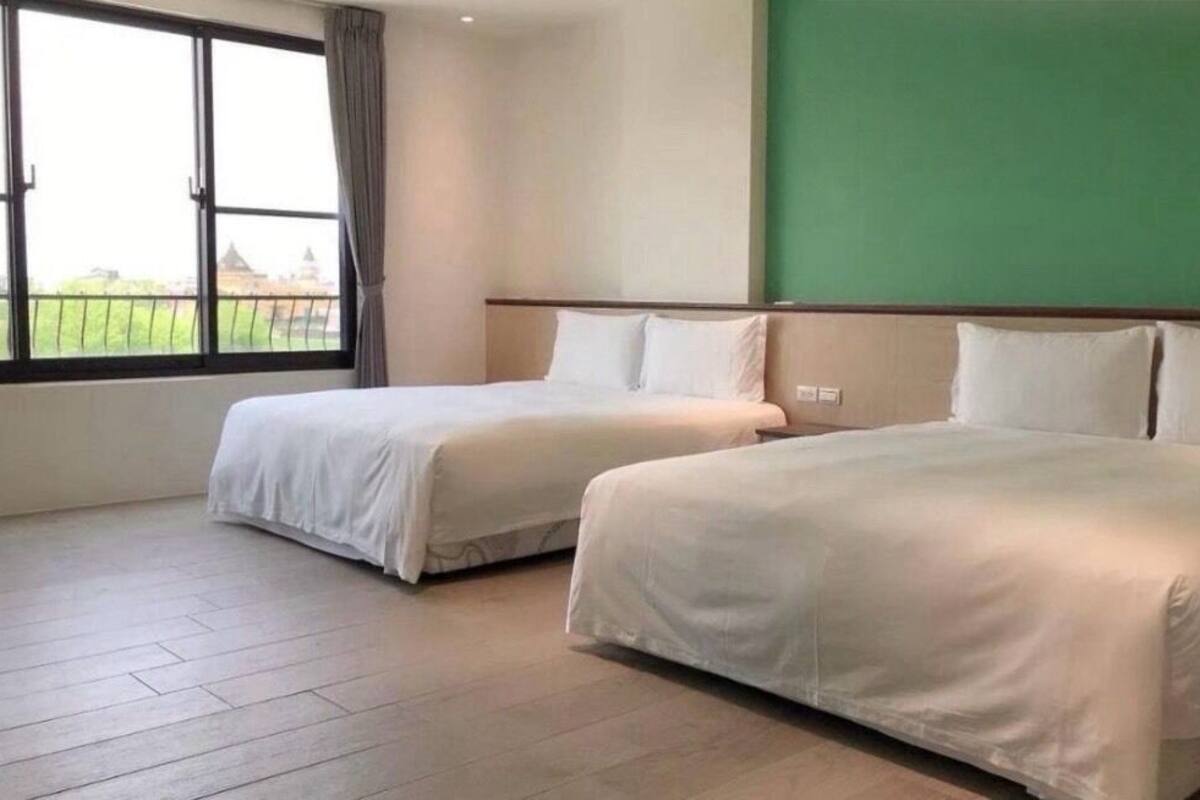 Quarto de hotel com duas camas, uma janela grande e uma parede verde decorativa.
