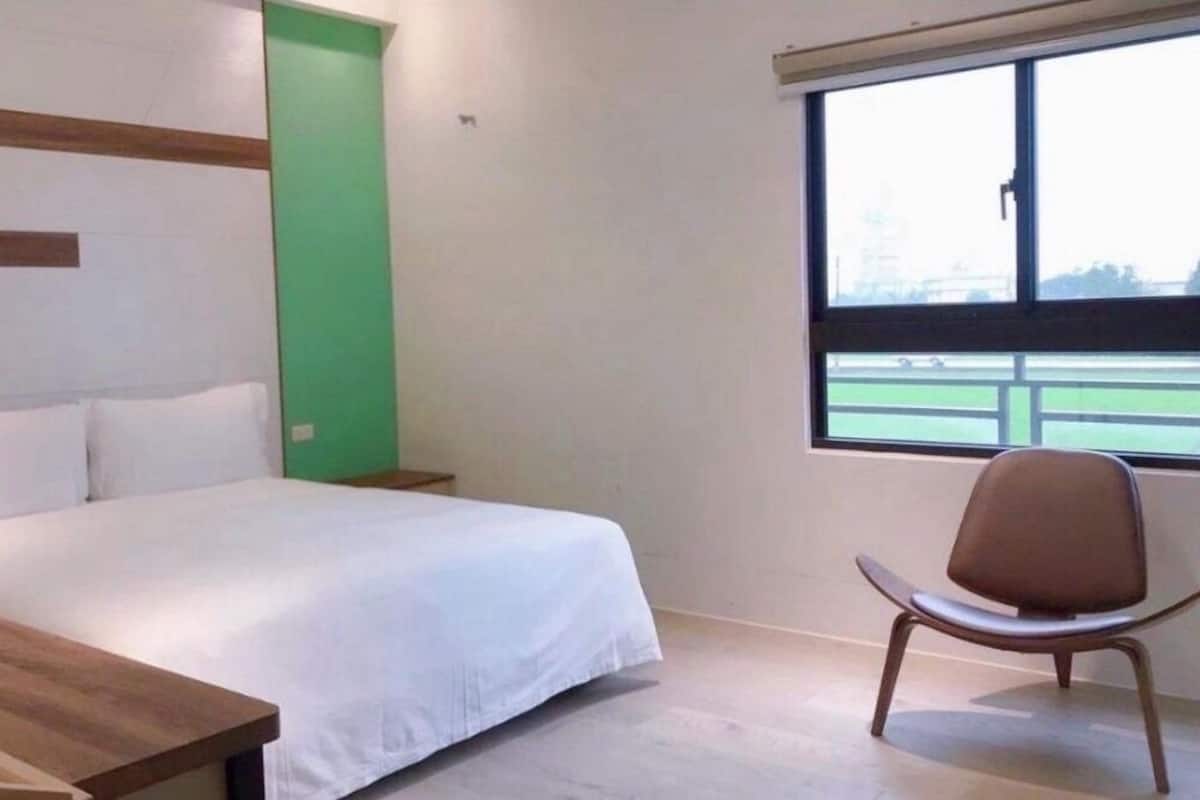 Quarto de hotel com cama, mesa de cabeceira de madeira, parede verde decorativa e janela com vista para um campo esportivo.