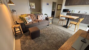 A modern living room with a sofa, a coffee table, and a rug. There is a dining area with a table and chairs. The kitchen is visible in the background.