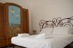 A neatly made bed with white linens, a wooden wardrobe with a colorful painting, and a metal headboard.