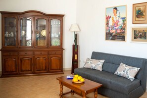 A living room with a wooden cabinet, a grey sofa, a table with fruit, and a lamp.