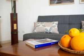 A book on a table, a basket of oranges, and a grey sofa with patterned cushions.