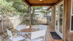 A covered outdoor patio with a wooden table and white chairs, surrounded by a wooden fence and trees. - Beachcomber Cottage · Close to Beach, Family Friendly, Open Layout (Pacific City)