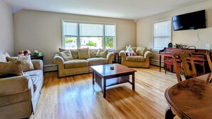 Ein Wohnzimmer mit Holzboden, zwei beigefarbenen Sofas, einem Couchtisch, einem Esstisch mit Stühlen und einem an der Wand befestigten Flachbildfernseher.