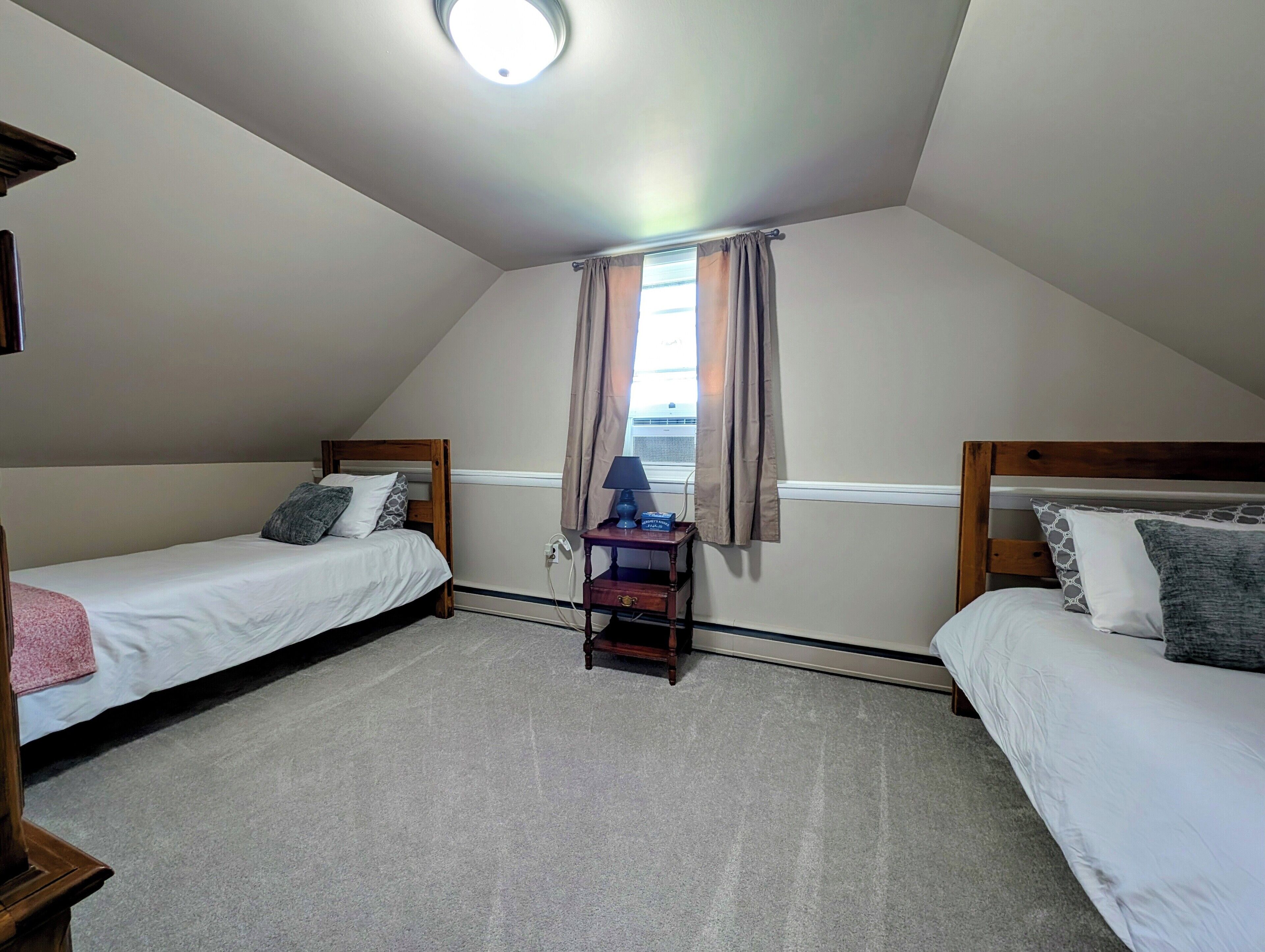 A small, well-lit attic bedroom with two single beds, a nightstand, and a window with curtains.