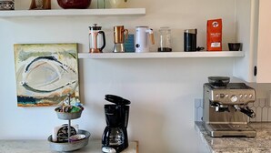 Eine Küche mit einer Kaffeemaschine, einem Mülleimer und einem Regal mit verschiedenen Küchenutensilien.
