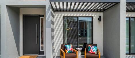 A modern building with a glass door entrance, a small outdoor seating area with two chairs and a table, and a covered patio area.