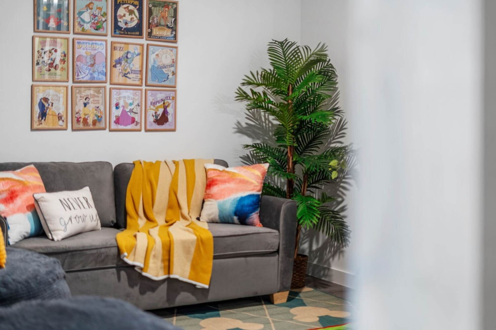 Ein Wohnzimmer mit grauem Sofa, bunten Kissen, einer gelben Decke und einer großen Grünpflanze.