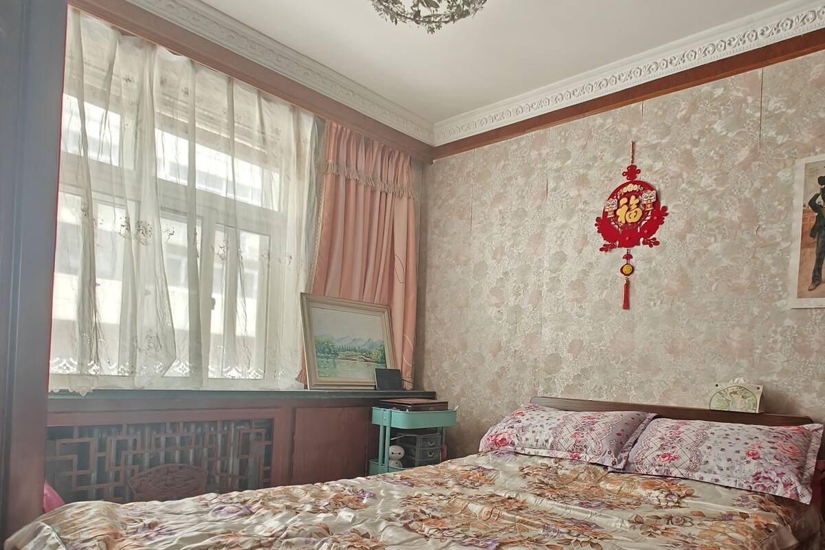 Ein Schlafzimmer mit blumigem Bettbezug, einem Holzkopfteil, einem Nachttisch, einem gerahmten Bild und einem dekorativen Wandornament.