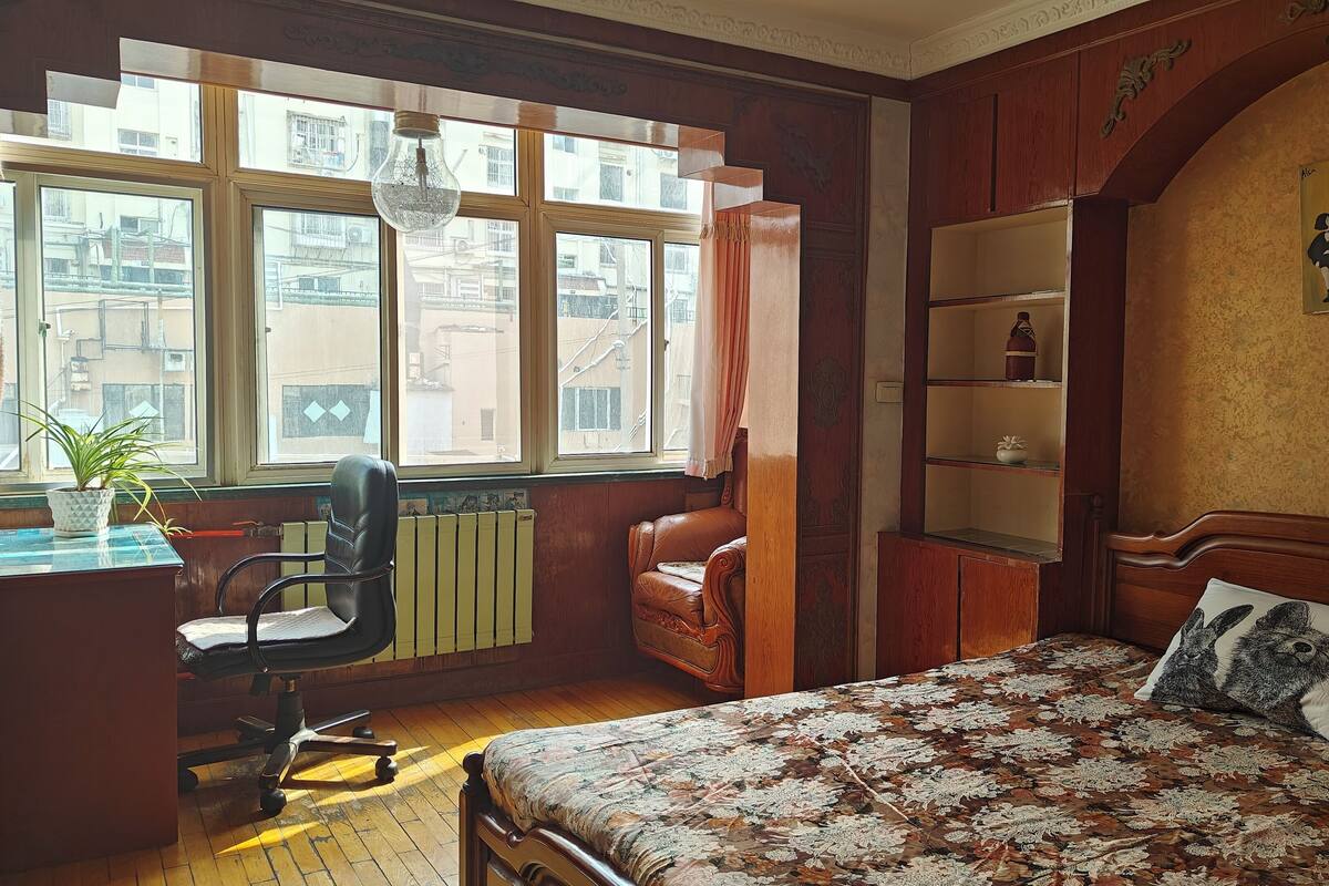 Ein Schlafzimmer mit großem Fenster, einem Schreibtisch mit Pflanze, einem Ledersessel, einem Bett mit floralem Bettbezug und einem Regal mit Dekoration.