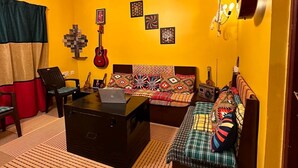 Una habitació amb una paret groga, un llit amb roba de llit estampada, un escriptori amb un ordinador portàtil, una guitarra i quadres emmarcados a la paret.