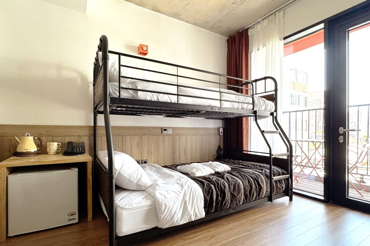 A bunk bed room with a small kitchenette, a window with curtains, and a door leading outside.