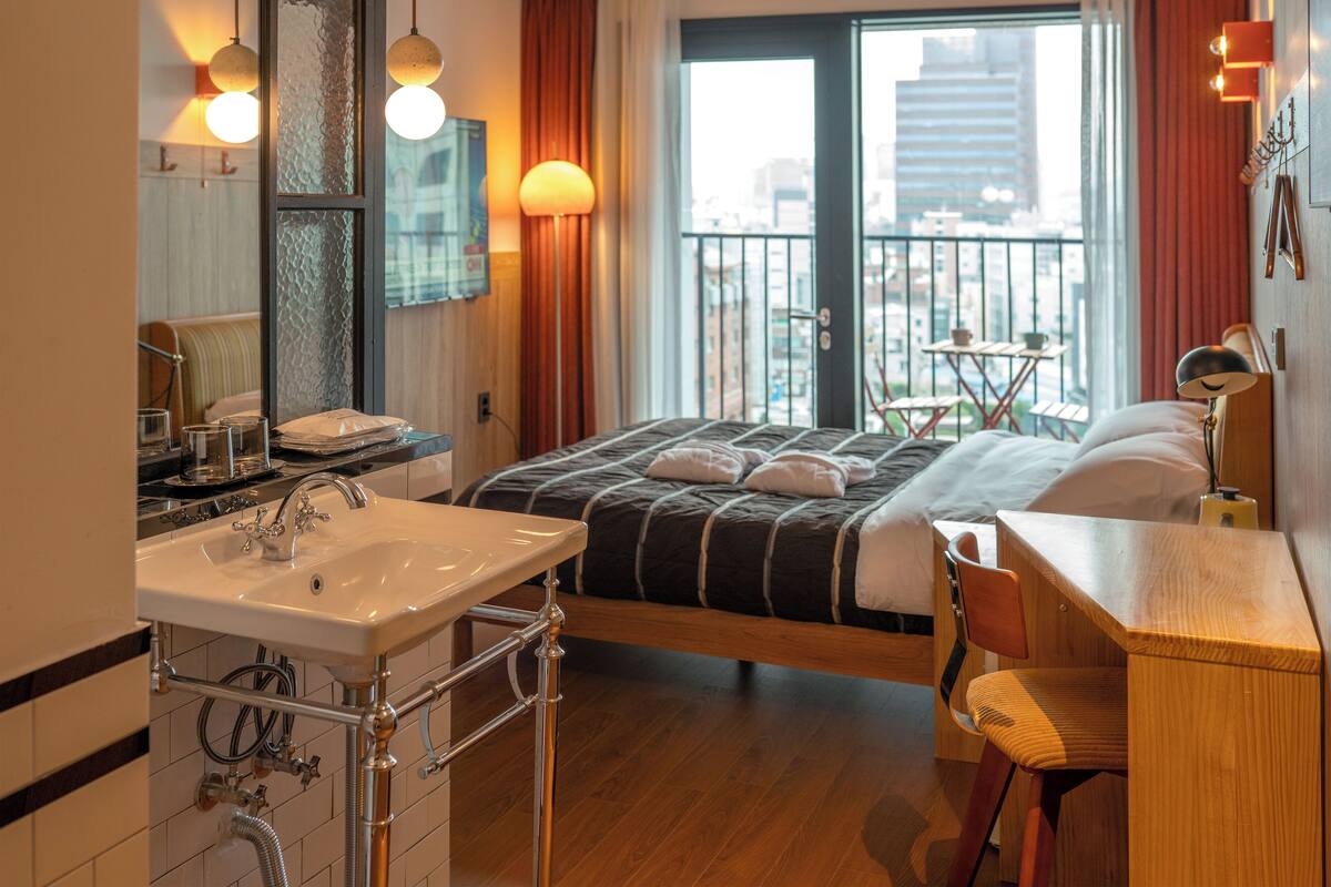 A hotel room with a large bed, a wooden desk, a bathroom with a sink and mirror, and a balcony with a view of the city.