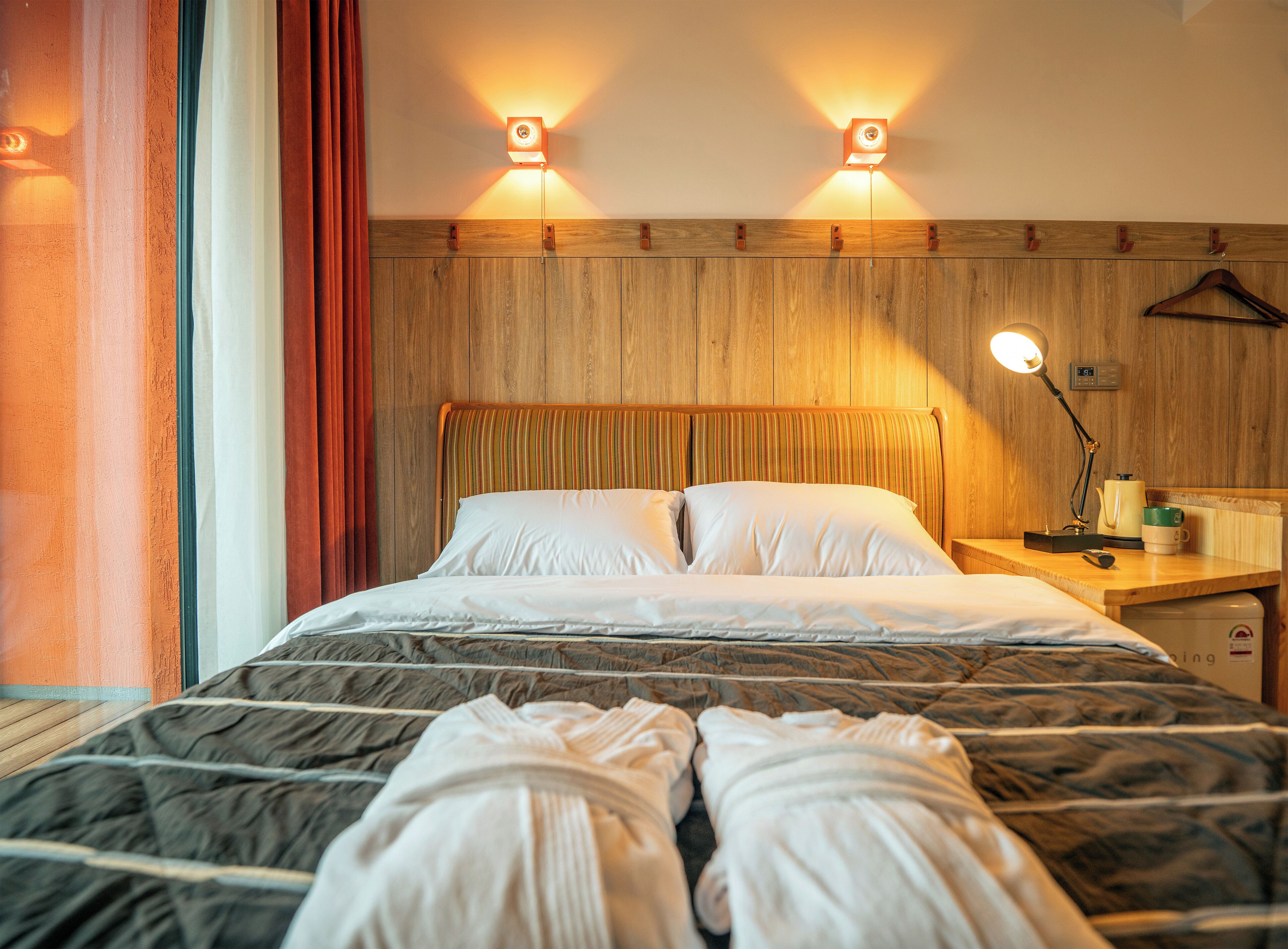 Una habitación de hotel con cabecera de madera, dos lámparas de noche, mesitas de noche de madera y una cama con ropa de cama blanca.