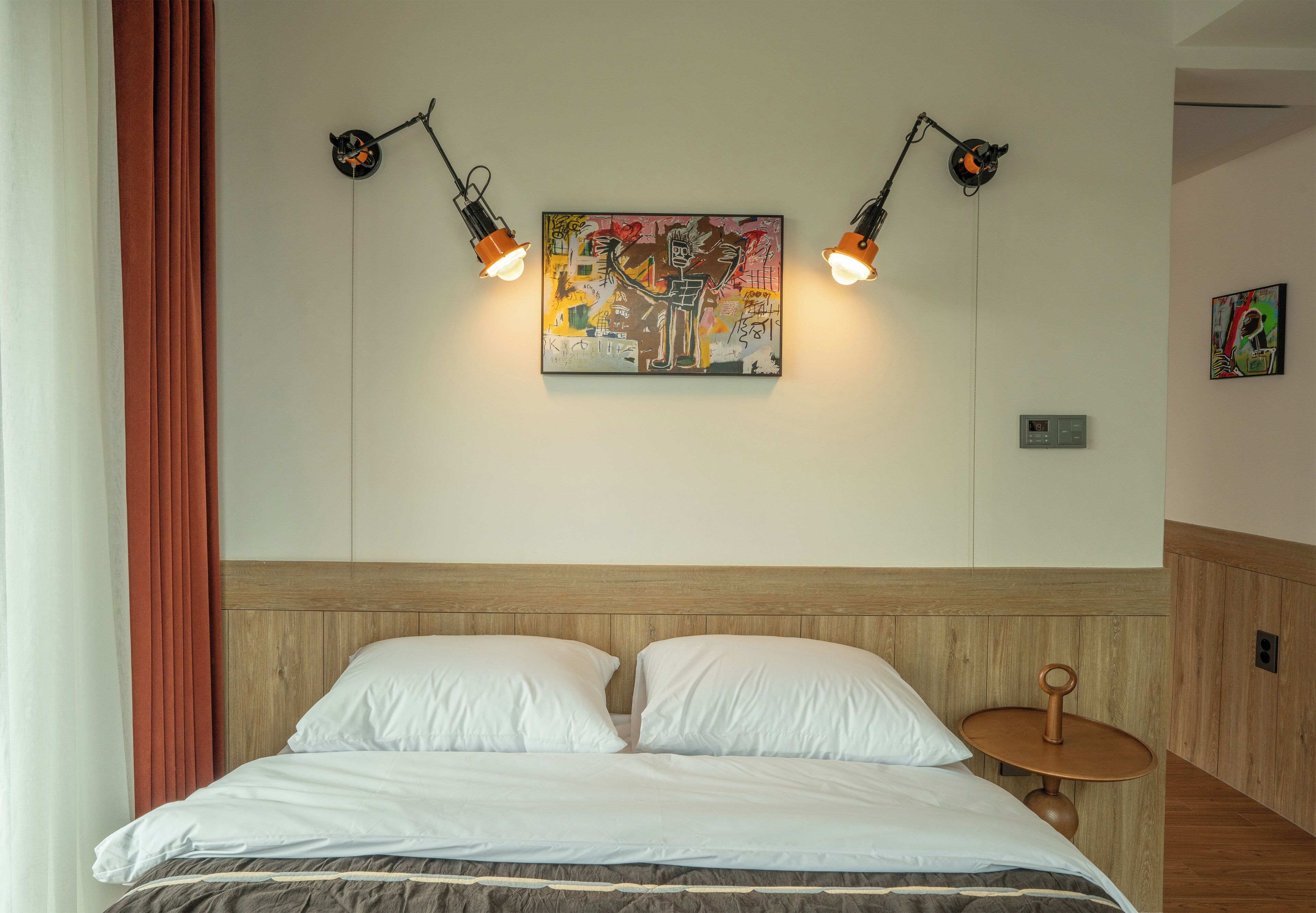 A neatly made bed with white linens, a wooden headboard, and a wall-mounted painting with two wall-mounted lights.