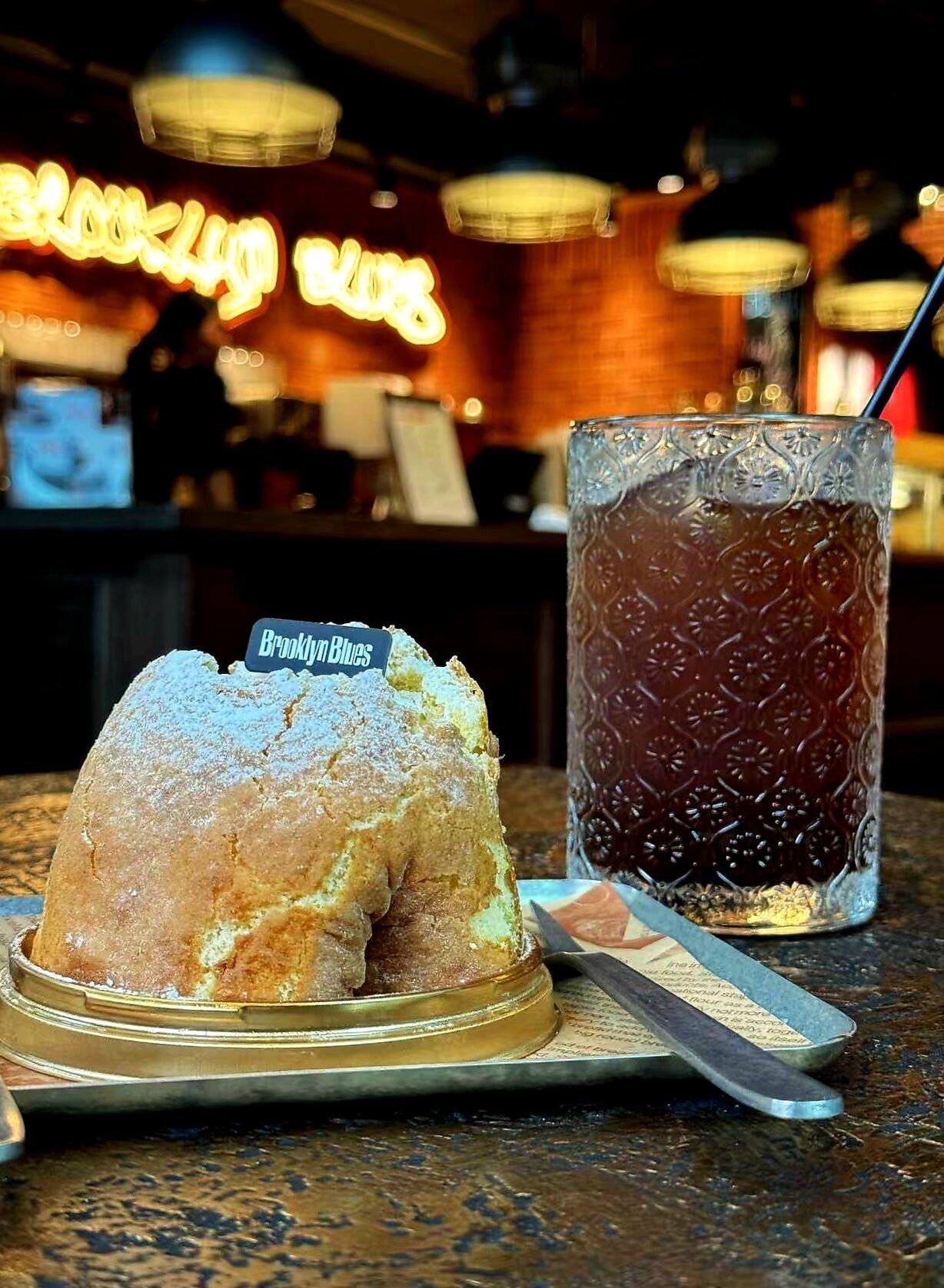 Un dessert présenté sur une assiette, accompagné d’une étiquette, un verre de boisson fraîche et un comptoir de bar en arrière-plan.