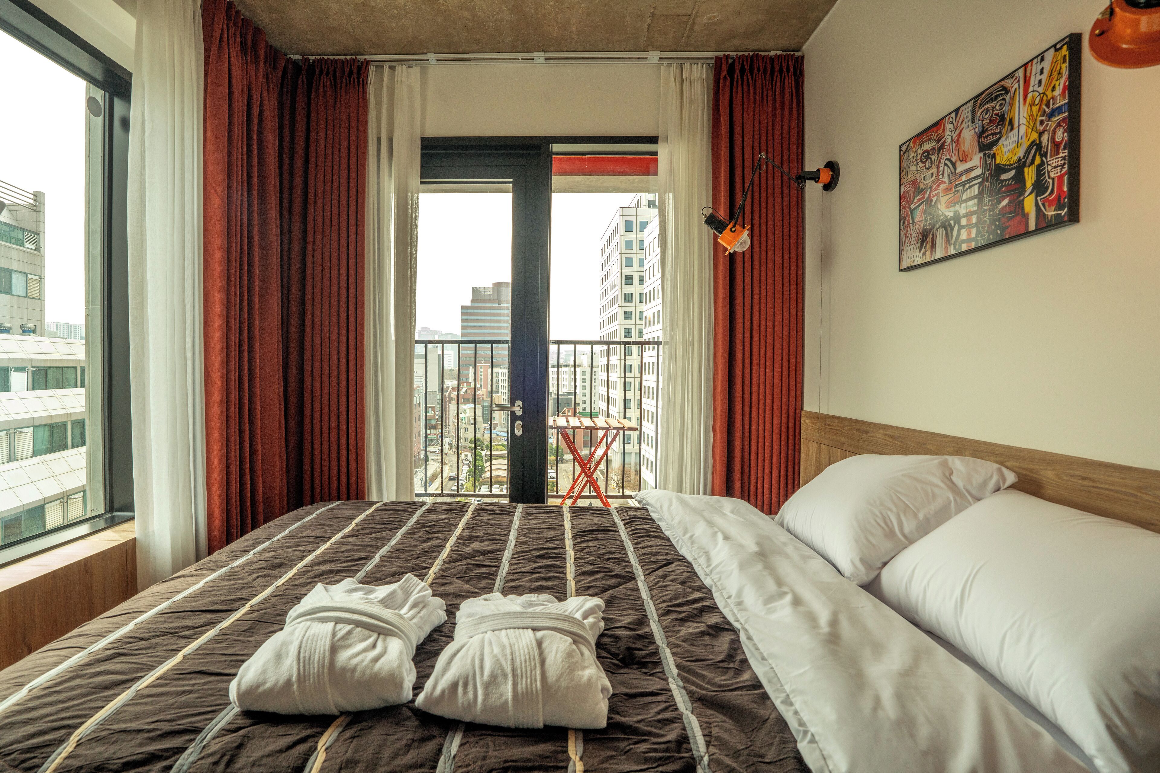 A hotel room with a bed, a view of the city, and a painting on the wall.