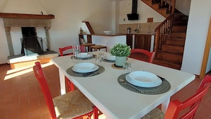 A dining area with a white table set for four, surrounded by red chairs with woven seats. A small potted plant is on the table, and a staircase is visible in the background.
