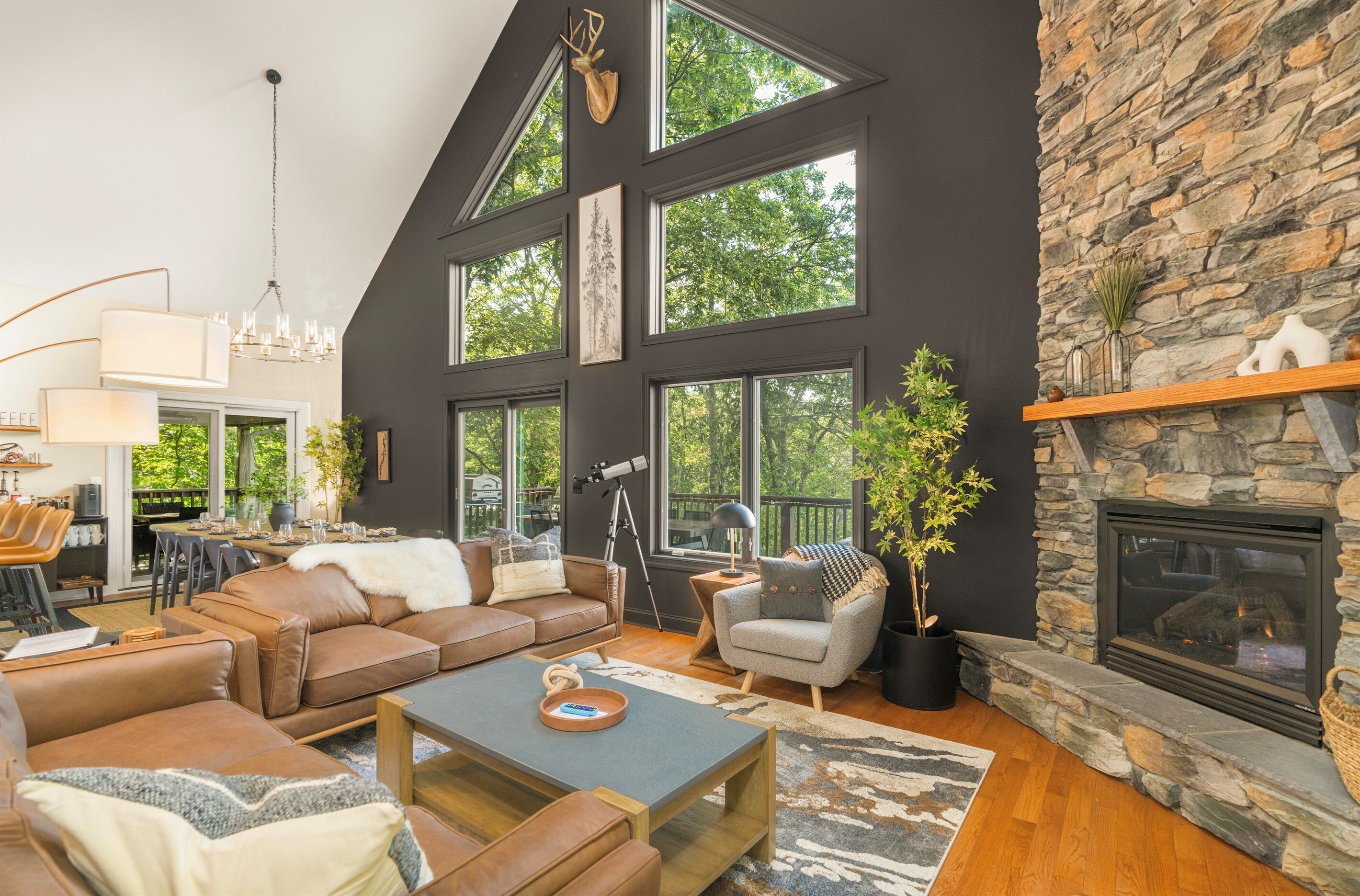 A spacious living room with leather sofas, a stone fireplace, large windows, and a dining area with a chandelier.