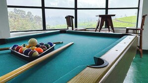 A pool table with balls and cues, a wooden chair, and a view of a green landscape through large windows. - Dover Cottage (Nuwara Eliya)