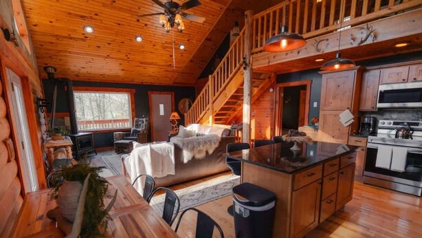 A rustic cabin interior with a wooden dining area, a cozy living room with a fireplace, a kitchen with modern appliances, and a staircase leading to an upper level. - COZY Cabin with Hot Tub - Near all in Hocking Hills (Logan)