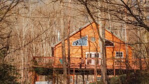 A wooden cabin with a balcony, surrounded by trees and a chair in the foreground. - COZY Cabin with Hot Tub - Near all in Hocking Hills (Logan)