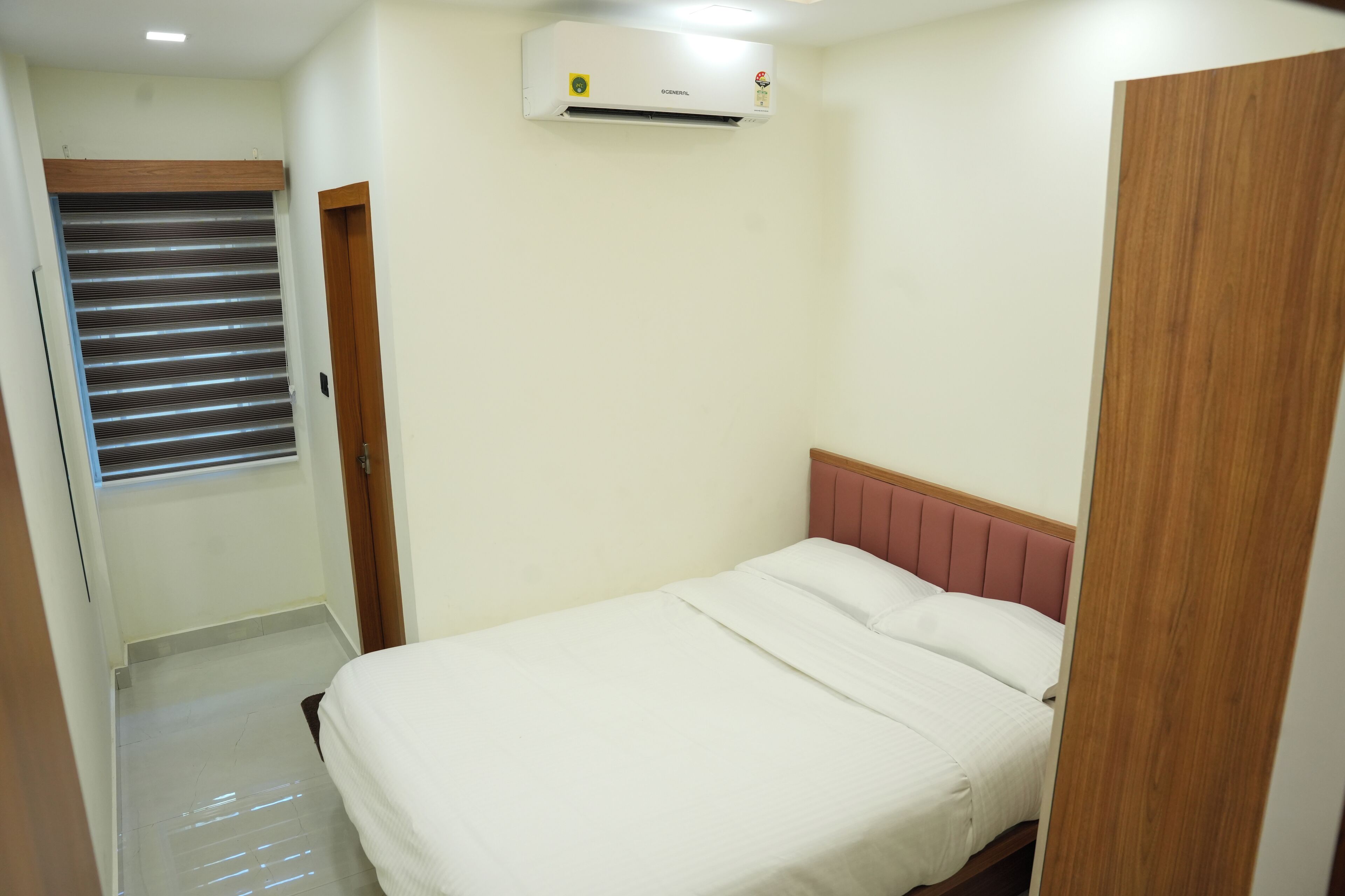 Ein Hotelzimmer mit einem Bett, einem Fenster mit Jalousien, einer Klimaanlage und einer Holztür.