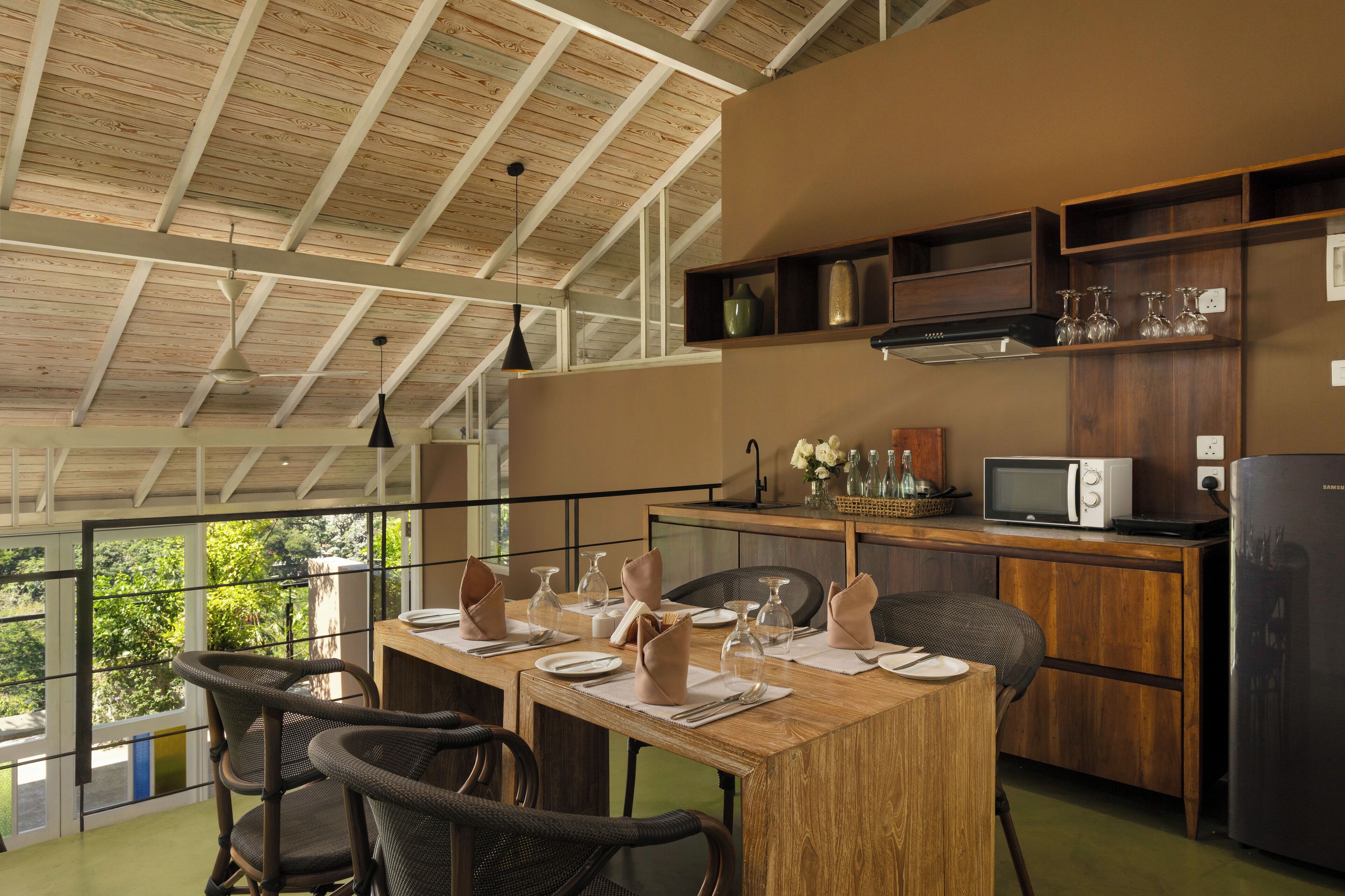 A dining area with a wooden table set for four, a microwave, a refrigerator, and a view of greenery outside.